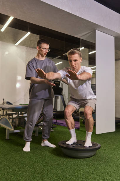 istock Man performing squats on balance trainer, receiving support and feedback from trainer during rehabilitation session. 2190685645