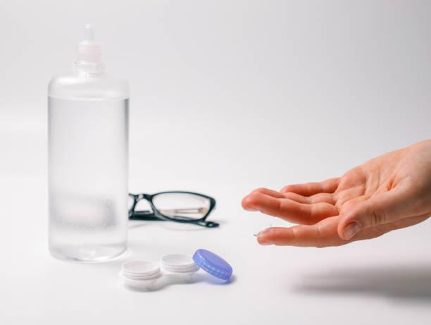 istock Hand holding a transparent contact lens next to lens case, solution bottle, and glasses on a white background. Concept of eye care, vision correction, and optical hygiene 2190691821