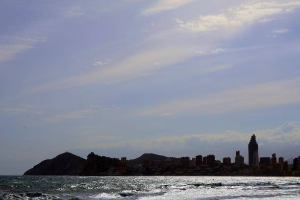 A stunning coastal landscape featuring gentle ocean waves lapping against the shore. The silhouette of a prominent city skyline rises in the background, with modern buildings reflecting the soft light of the setting sun. To one side, a scenic hillside adds depth to the view, creating a serene atmosphere perfect for relaxation and contemplation.