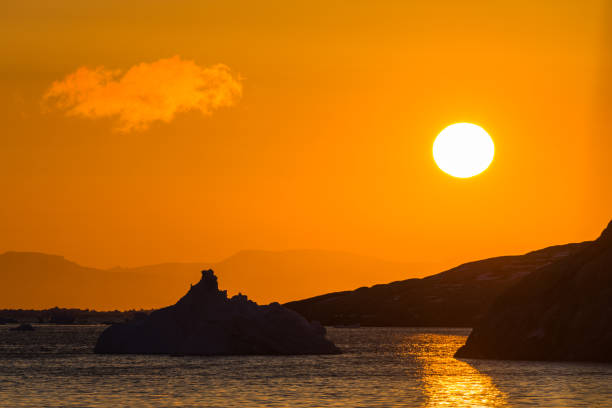 A mountain range in a Greenland Fjord is seen at early sunrise.  An iceberg is partially lit in the photo.  The colors range from orange to blue.