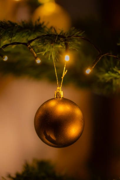 A gold colored Christmas ornament hangs from a tree. The ornament is a ball and is hanging from a string