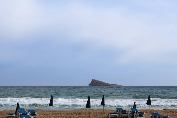 This tranquil beach scene features rows of blue lounge chairs under dark umbrellas, inviting relaxation along a golden sandy shore. In the distance, a distinctive rocky island juts out from the turquoise waters, framed by gentle waves. The cloudy sky creates a serene atmosphere, perfect for evoking the essence of a peaceful getaway by the sea.