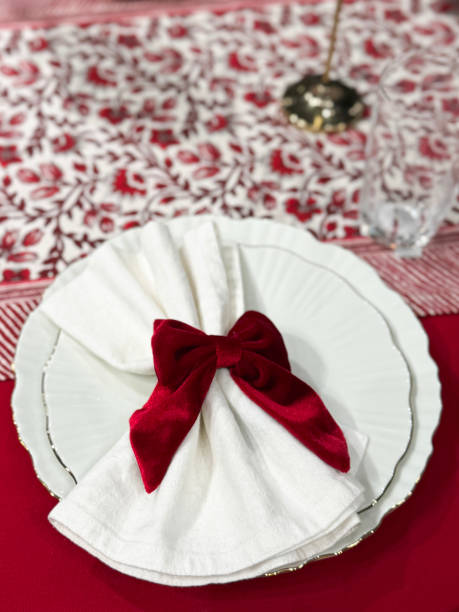 istock Close-up image of Christmas dining table place setting, stacked scalloped edge, dinner and tea plates, white napkin with red velvet bow design napkin ring, drinking tumblers, red tablecloth and table runner, elevated view 2190697945