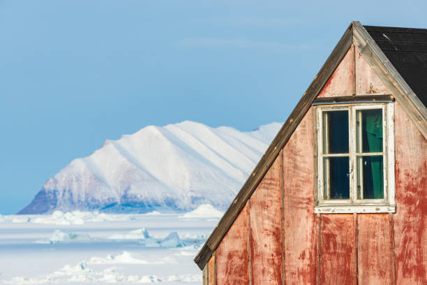 A rustic red cabin stands in the foreground with a window reflecting light. In the background, majestic snow-capped mountains rise against a clear blue sky, showcasing the serene winter beauty.