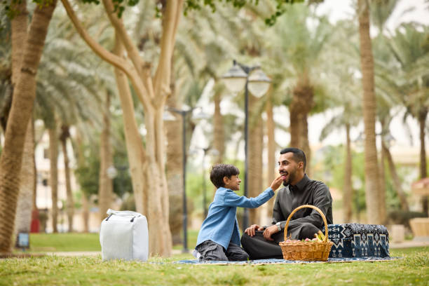 Son feeding fresh fruits to father sitting on blanket during picnic. Boy is spending quality time with parent. They are at garden.