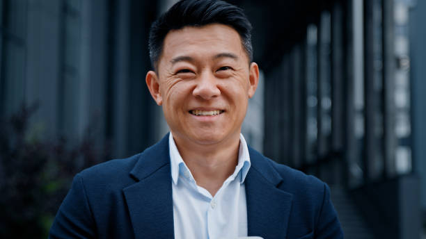 istock Glad Asian 40s businessman with smartphone texting browsing smiling looking at camera standing outdoors in city. Middle-aged man CEO has business chat using mobile phone check news in social net 2190713931