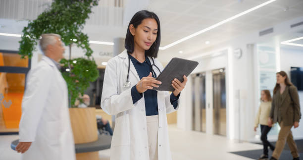 istock Portrait of an Asian Female General Practitioner Using Tablet Computer to as She is Browsing Insights About a Complex Medical Case. Healthcare Workers Walking Through a Modern Hospital Hall 2190715406