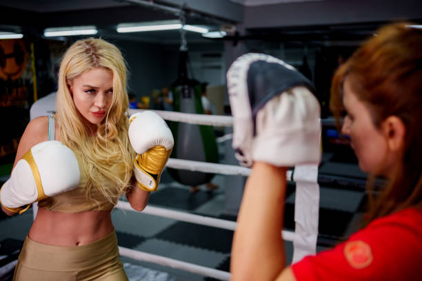 A young blonde woman, wearing white boxing gloves and a focused expression, practices her boxing stance with a trainer holding punch pads in a gym. The girl appears determined and attentive as she learns boxing techniques, representing strength, discipline, and empowerment in youth sports.