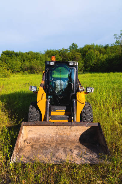 red backhoe with large wheels parked on edge of dusty road under cloudy sky, surrounded by grass and trees