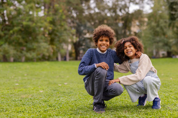 In a peaceful park setting, a little black brother and his sister looking at camera while sharing a playful moment together. Their bright smiles and the natural, green surroundings create an atmosphere of happiness and sibling connection in this candid