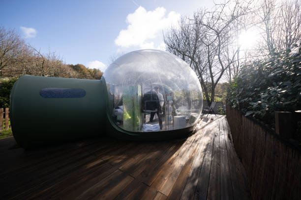istock Couple standing inside transparent bubble hotel room, connected to green cylindrical structure, relaxing on wooden terrace during glamping experience 2190738256