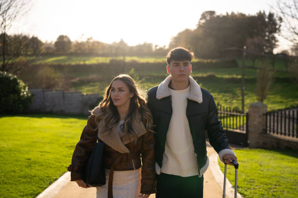 istock Young couple walking countryside path, pulling rolling suitcase, wearing stylish jackets, enjoying serene rural landscape during leisurely travel moment 2190738336