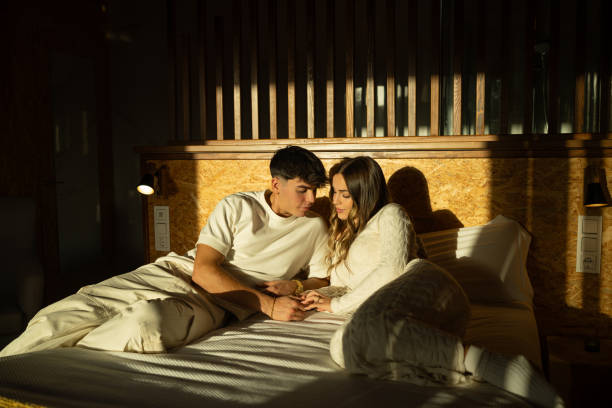 istock Young couple sitting intimately on bed, holding hands, sharing tender moment while soft morning sunlight streams gently across their embracing figures 2190738461