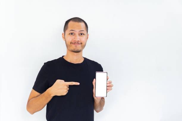 istock Young men with black empty shirt  smiling and pointing to phone with white screen template mockup 2190766816