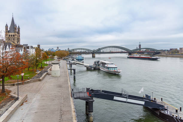 The Rhine River waterfront in Cologne, boats and barges anchored for loading, railway bridge over the river in the background under the cloudy sky, residential area with church near promenade