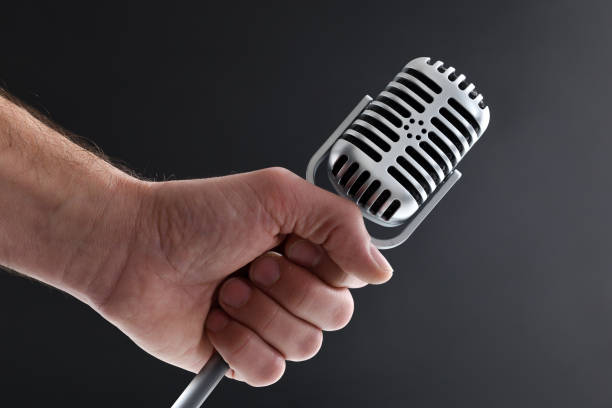 Classical musical vocal performance concept with hand holding a vintage style metal condenser microphone with stand on a dark isolated background. Front view.