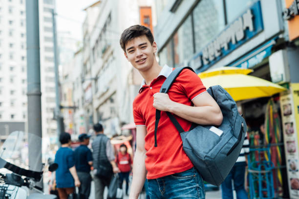 istock Young Asian man in a red shirt with a backpack smiles while standing on a bustling city street with shops in the background 2190799637