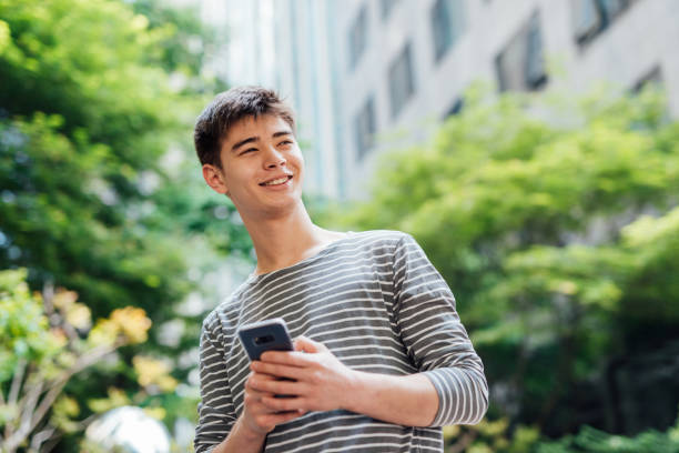 istock Young Asian man in a striped shirt smiling while texting on a smartphone outdoors with green foliage in the background 2190799750