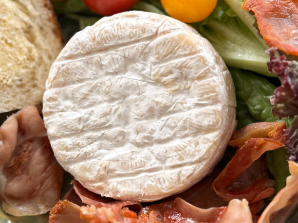 Stock photo showing close-up, elevated view of a whole brie cheese, prosciutto, lettuce leaf salad, red and orange cherry tomatoes, and white bread and butter.