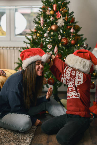 istock Little Boy Playing With Aunt's Santa Hat While Sitting in Front of Christmas Tree 2190809542