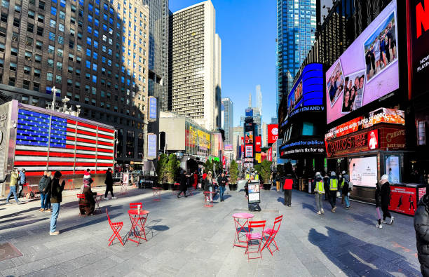 New York, NY, USA - December 13, 2024: Crowds stroll through Times Square amidst colorful advertisements and an American flag mural under a clear blue sky in New York City.
