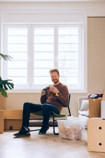 istock Man Relaxing in Modern Home with Smartphone Amid Moving Boxes 2190846951
