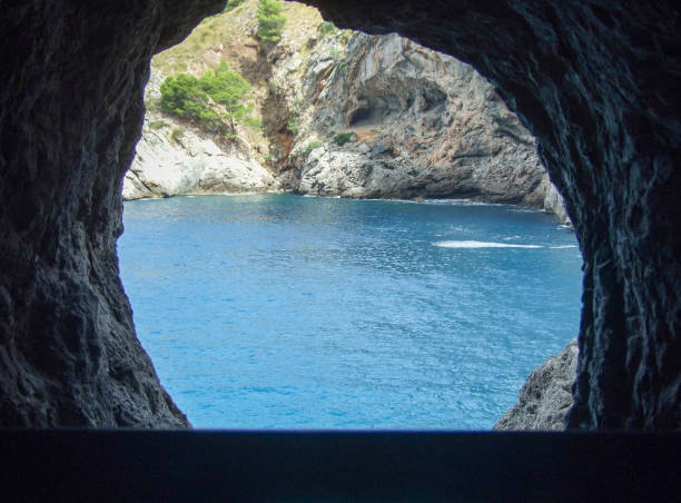 Through a natural rock formation, a stunning view reveals clear blue waters and vibrant coastal cliffs. The sunlight reflects off the calm sea, creating a serene atmosphere in this secluded spot.