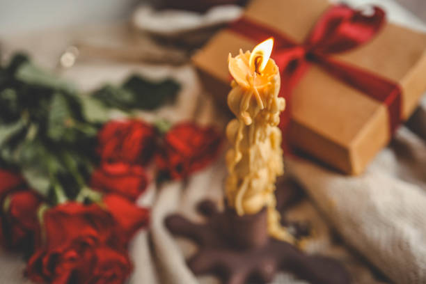 istock A cozy window-side scene featuring a lit beeswax candle with a unique shape, vibrant red roses, a gift box with a ribbon, and a soft textured fabric, creating a warm and serene ambiance 2190851991