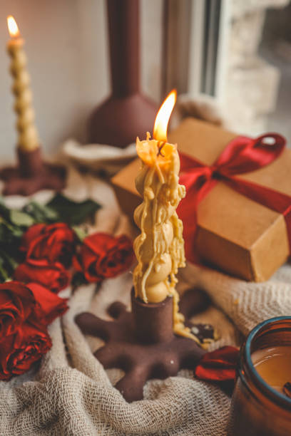 istock A cozy window-side scene featuring a lit beeswax candle with a unique shape, vibrant red roses, a gift box with a ribbon, and a soft textured fabric, creating a warm and serene ambiance 2190851993