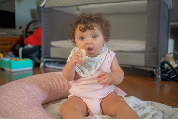 A baby with curly hair, wearing a soft bib, sitting on a play mat and enjoying a piece of food. A natural moment capturing playtime and exploration at home