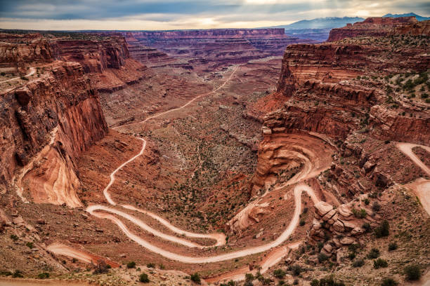 The image shows the Shafer Trail, a winding dirt path through a scenic desert canyon. The rugged terrain features contrasting rock formations and natural colors, effectively showcasing the beauty of the landscape.