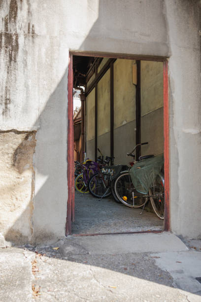 Pointed shadows stretch across a building's rough, concrete wall, creating an interesting contrast of light that covers a doorway opening into an area with parked bicycles. The image showcases everyday urban life in Japan.