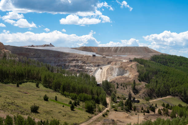 A vast mining operation unfolds within a mountainous region, with towering earth mounds and machinery visible. Lush green trees grow at the perimeter, highlighting the contrast between nature and industrial activity under a bright, cloudy sky.