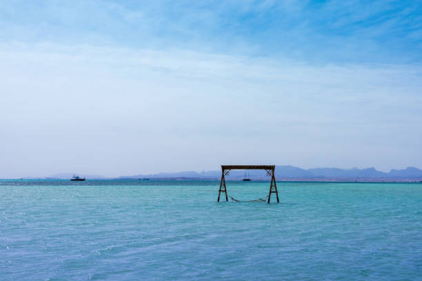 This serene image captures a tranquil seascape with a hammock gently suspended over calm blue waters, set against a backdrop of clear azure skies. the distant horizon and a solitary boat evoke a sense of peace and escapism, ideal for travel and leisure themes. with its soothing colors and minimalistic composition, this scene is perfect for conveying relaxation, nature, and the beauty of coastal life.
