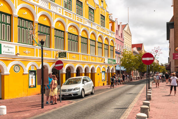 Willemstad, Curacao, Netherlands - January 6, 2018: A vibrant street scene in the historic district of Willemstad, featuring colorful colonial architecture, tourists, and a car parked along the cobblestone road.