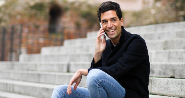 istock Young man sitting on stairs and talking on smartphone, smiling and enjoying conversation 2190907929