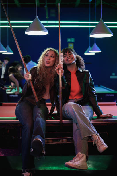 Two young women are making funny faces while sitting on the edge of a pool table, holding pool cues