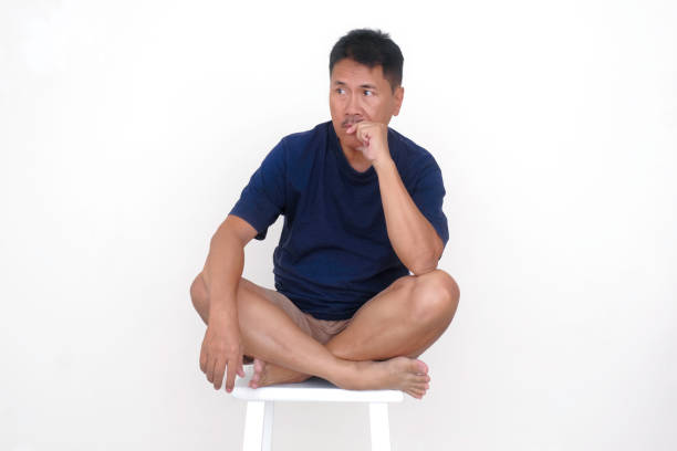 Asian guy in casual clothes, sitting alone on white stool thinking