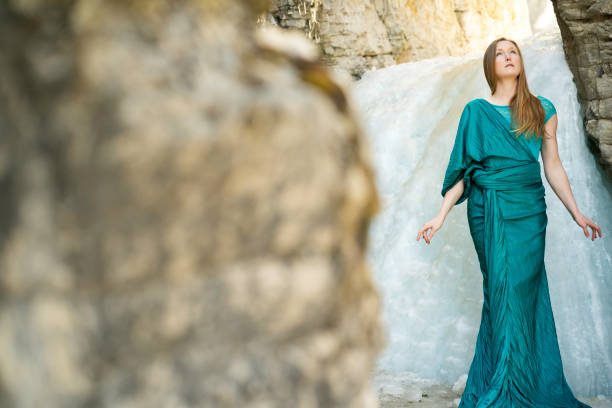Elegant woman with long auburn hair wearing a long turquoise dress stands near a frozen waterfall looking up in a canyon, Jasper, Alberta, Canada