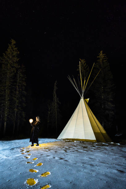 Woman holds an illuminated globe in her hands beside a teepee with yellow illuminated foot tracks in the snow, Jasper, Alberta, Canada