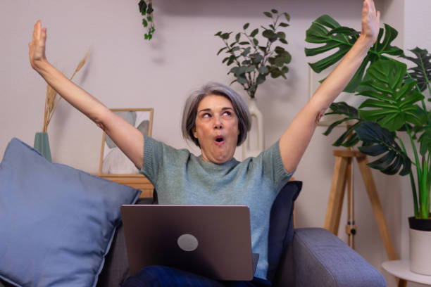 istock Excited mature woman raising arms while using laptop at home 2190939500