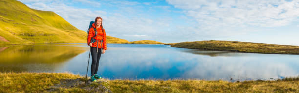 Teenage girl hiker wearing backpack and using hiking pole walking across the green mountain landscape of the Brecon Beacons National Park, Wales.