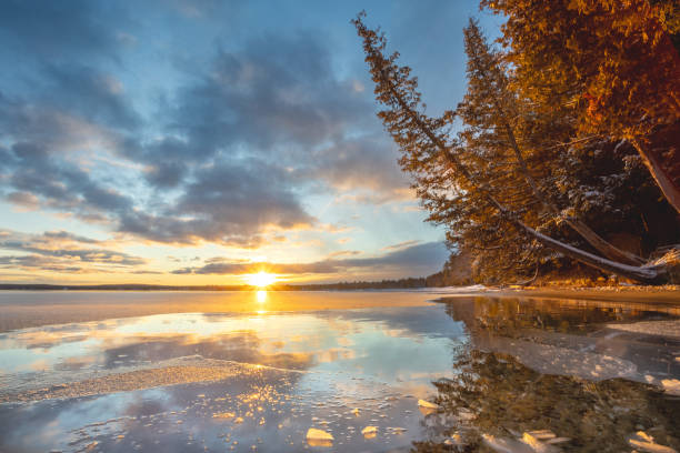A serene winter solace sunset at Higgins Lake Michigan casts a warm glow over a partially frozen lake, with the sky reflecting on the icy surface. Snow-dusted trees line the shore, adding a peaceful, natural ambiance to the scene. Higgins Lake North State Park, Michigan. 12/20/24
