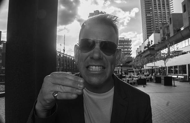 istock 40s Man in white t-shirt, suit jacket and aviators grits his teeth angrily to camera while gesturing with his signet ring hand. Cloudscape and central tower of Barbican estate behind him along with Barbican centre and it's central public court. 2190965902