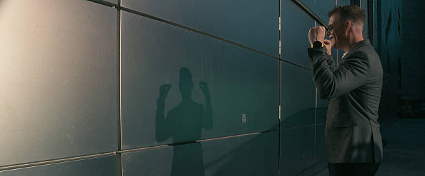 istock Man in white t-shirt, suit jacket, and aviators stands in profile, smiling, facing his shadow on a wall made from shiny steel panels - lit up by the golden setting sun. He makes 'chat' gestures with his hands either side of his head echoed by his shadow. 2190965919