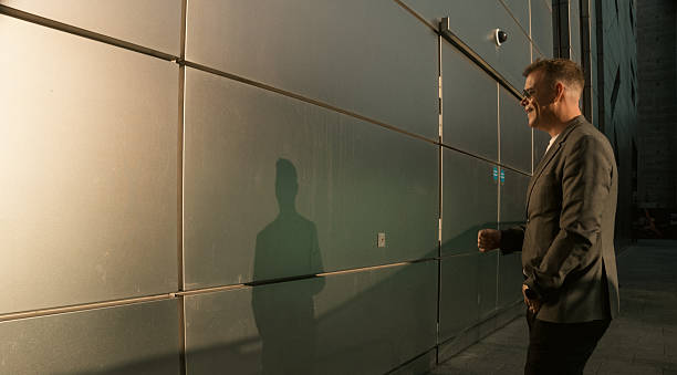 Man in white t-shirt, suit jacket, and aviators stands in profile, smiling, facing his shadow on an exteror office wall made from shining steel panels - lit up by the golden setting sun.