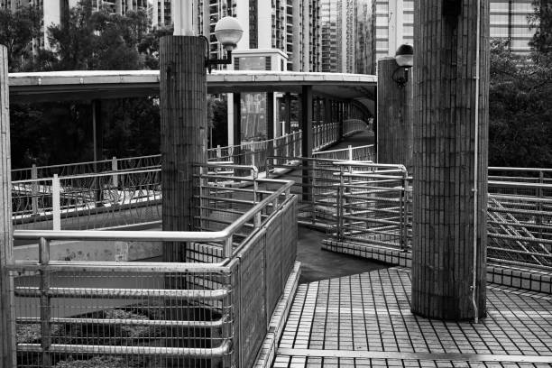 A black and white photograph of a modern urban walkway featuring metal railings and concrete pillars. The scene captures the intricate design of the pathway with a backdrop of tall buildings.