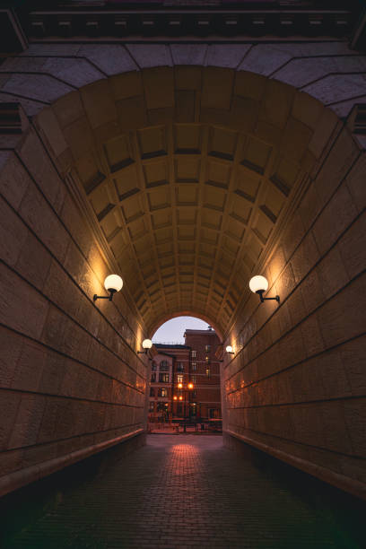 An illuminated urban archway at dusk with glowing streetlights and a city view beyond. The warm lighting and architectural details create an atmospheric and scenic urban scene