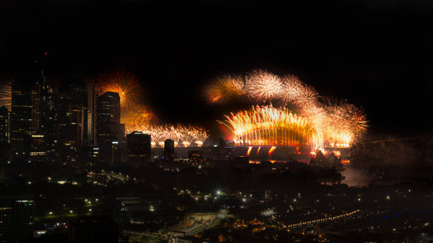Dramatic view of Sydney's modern skyline with illuminated skyscrapers framing the New Year's Eve fireworks display over Harbour Bridge and Opera House. Golden pyrotechnics reflect against glass buildings while city lights twinkle below, showcasing the scale of the celebration.