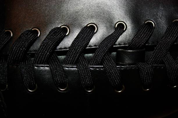 Detailed close-up of black leather shoe upper featuring black textile laces threaded through metallic eyelets. The surface texture exhibits a smooth finish and subtle sheen.